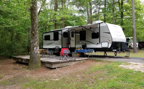 A parked RV in a wooded campsite, with a wooden deck and outdoor seating set up nearby.