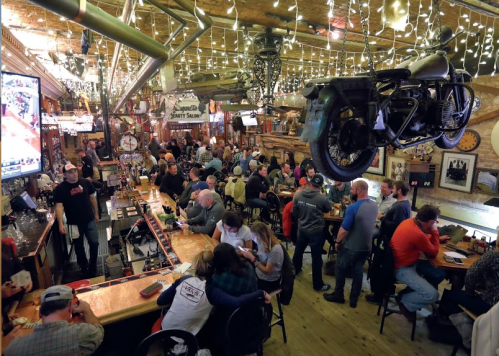 A lively bar scene with patrons enjoying drinks, decorated with string lights and a motorcycle hanging from the ceiling.