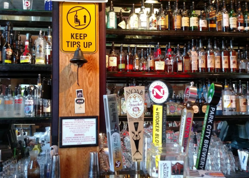 A bar counter with various beer taps and a shelf filled with liquor bottles, featuring a "Keep Tips Up" sign.
