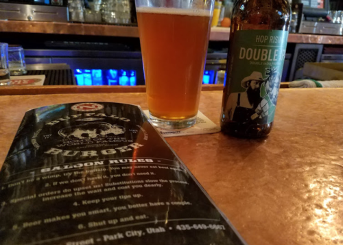 A glass of amber beer next to a bottle labeled "Hop Rising Doubler" and a menu on a bar counter.