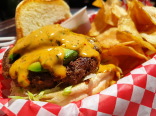 A juicy burger topped with melted cheese and green peppers, served with crispy potato chips on a red-checkered plate.