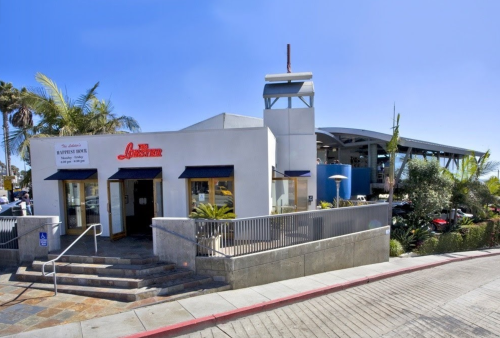 A modern restaurant with a white exterior, palm trees, and outdoor seating, set against a clear blue sky.