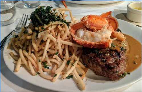 A plate featuring a grilled steak topped with lobster and a side of crispy French fries, garnished with herbs.