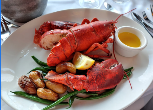A plate featuring a whole lobster, lemon, green beans, and roasted potatoes, served with a small dish of olive oil.