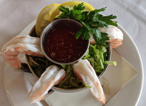 A bowl of shrimp on a bed of lettuce, garnished with lemon slices and parsley, served with a side of cocktail sauce.