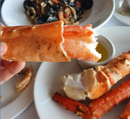 A hand holding a piece of cooked crab leg, with a plate of seafood in the background.