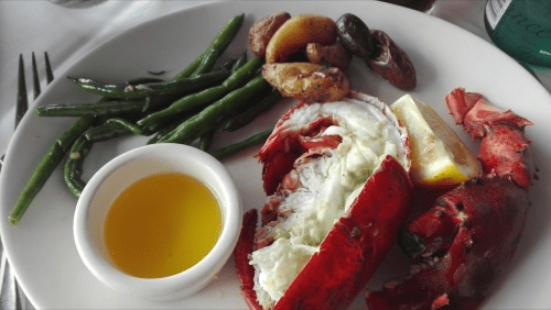 A plate featuring lobster with lemon, green beans, roasted potatoes, and a small dish of olive oil.