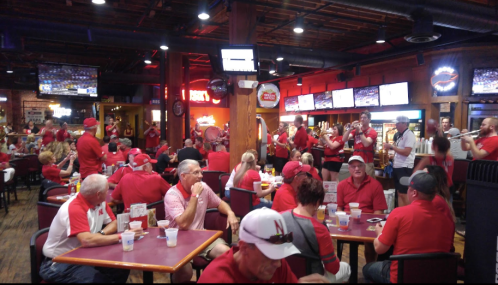 A lively restaurant filled with people in red attire, watching sports on multiple screens and enjoying food and drinks.