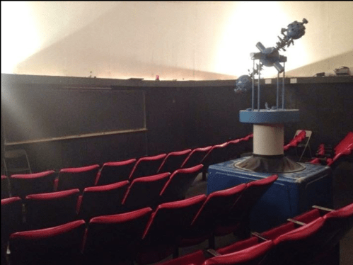 A small theater with red seats and a central planetarium projector under dim lighting.