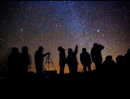 Silhouettes of people stargazing under a starry night sky, with a telescope set up in the foreground.