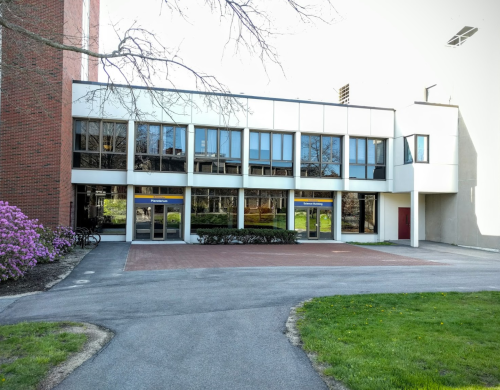 Modern building entrance with large windows, surrounded by greenery and blooming flowers, on a sunny day.