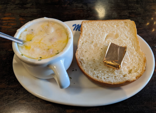 A cup of creamy soup with vegetables, served alongside a slice of bread and a butter packet on a white plate.