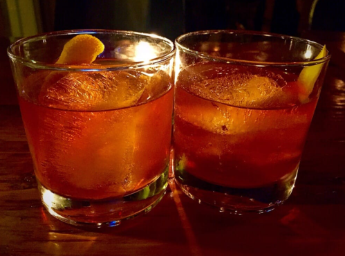 Two glasses of dark cocktails with ice and orange peel, illuminated by candlelight on a wooden table.