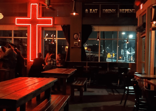 A dimly lit bar with a large red cross neon sign and a sign that reads "EAT DRINK REPENT." Tables are empty.