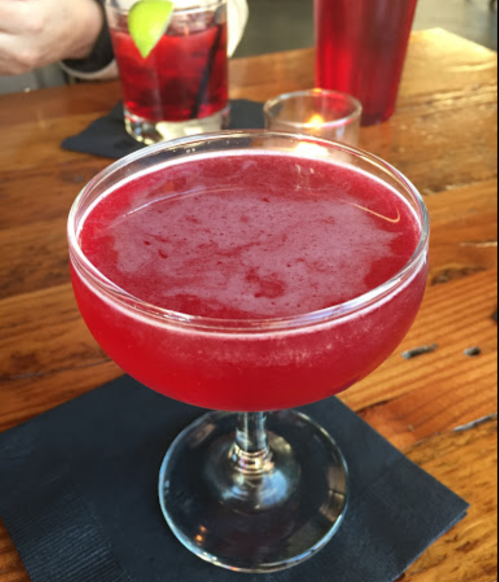 A vibrant red cocktail in a coupe glass, sitting on a wooden table with a candle and drinks in the background.