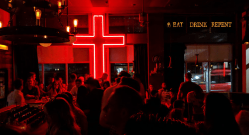 A lively bar scene with a large neon cross and a sign reading "EAT DRINK REPENT" in a dimly lit atmosphere.