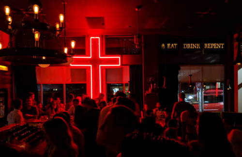 A lively bar scene with a large red neon cross and the words "EAT DRINK REPENT" illuminated in the background.