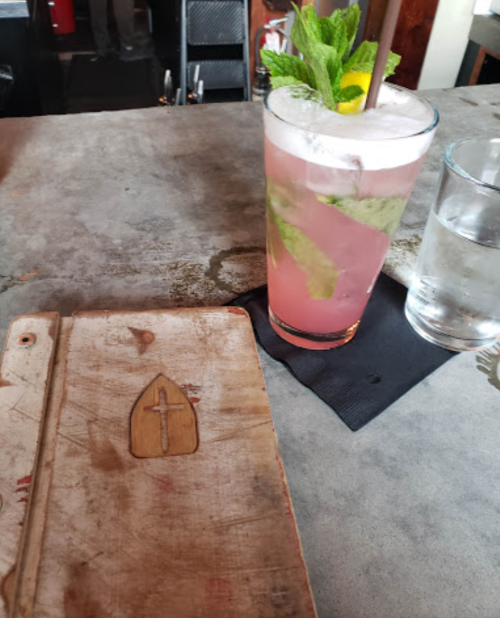 A wooden notebook with a cross design next to a pink cocktail garnished with mint and a lemon slice on a table.