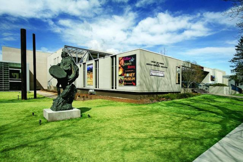 A modern museum building with a green lawn and a sculpture in front, under a blue sky with clouds.