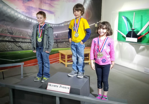 Three children stand on an Olympic podium, each wearing medals and smiling, in a sports-themed setting.