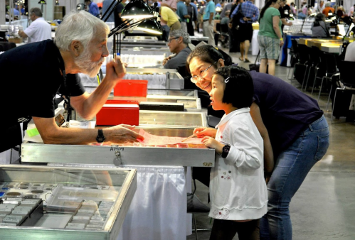A man and a woman engage with a young girl at a display table in a busy exhibition hall.