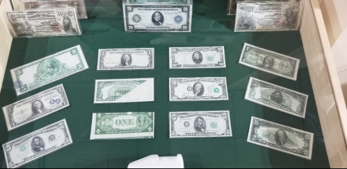 A display case featuring various U.S. banknotes, including $1, $5, $10, $20, and $100 bills, arranged on a green background.