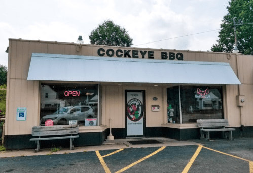 Exterior of Cockeye BBQ restaurant with an "Open" sign and benches outside, set against a cloudy sky.
