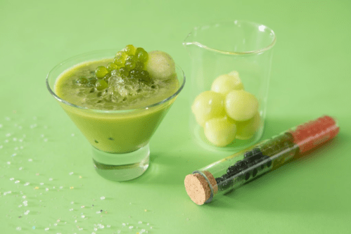 A green smoothie in a glass, topped with melon balls and green caviar, alongside a test tube filled with colorful beads.