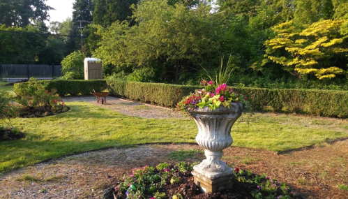 A garden scene featuring a large flower pot, manicured hedges, and a portable restroom in the background.