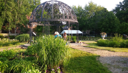 A decorative gazebo surrounded by lush greenery and flowers, with people tending to the garden nearby.