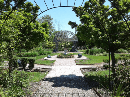 A serene garden with a stone gazebo, surrounded by lush greenery and a gravel path leading to the center.