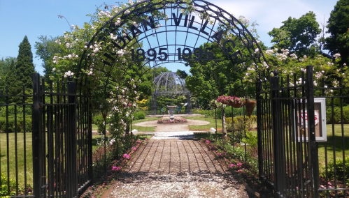 A wrought iron gate adorned with flowers leads into a lush garden with a fountain and a gazebo in the background.