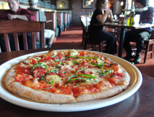 A freshly baked pizza topped with tomatoes, cheese, and basil, served in a restaurant with diners in the background.
