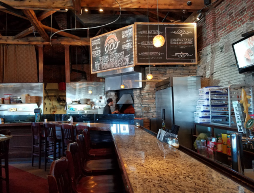 A rustic restaurant interior featuring a bar, wooden beams, and a chalkboard menu, with a chef visible in the kitchen.