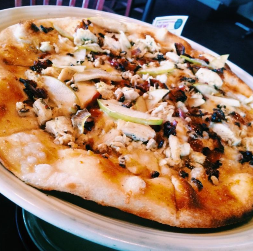 A close-up of a pizza topped with chicken, green apple slices, and herbs on a golden crust, served on a plate.