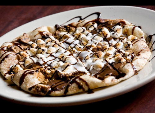 A dessert pizza topped with chocolate drizzle, marshmallows, and graham cracker crumbs on a white plate.