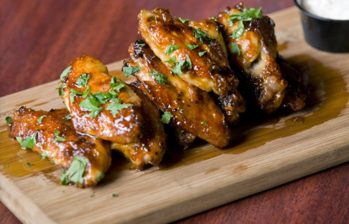 A stack of glazed chicken wings garnished with fresh herbs on a wooden serving board, accompanied by a dipping sauce.