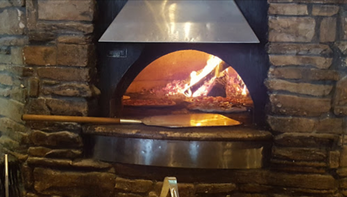 A stone pizza oven with a wooden pizza peel resting on the edge, flames visible inside.