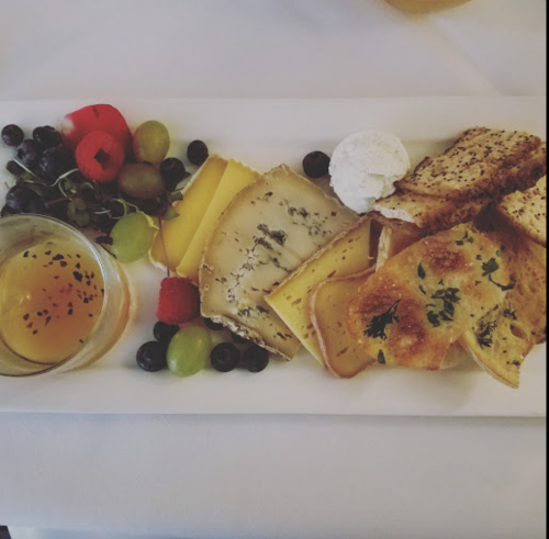 A cheese platter featuring various cheeses, fruits, a dollop of cream, and a small dish of honey.