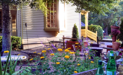 A sunny garden with colorful flowers, tables, and chairs beside a charming house. Greenery and potted plants are visible.