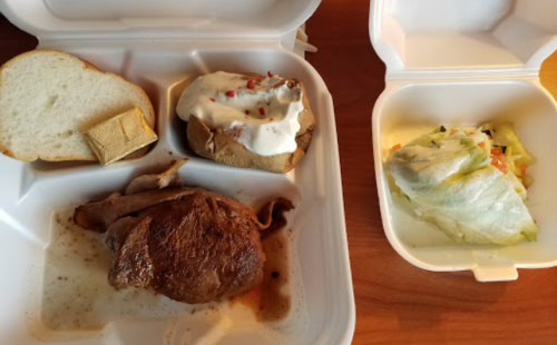 A takeout meal featuring a slice of bread, baked potato with sour cream, sliced meat, and a side salad.
