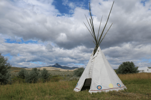 A white teepee with wooden poles stands in a grassy field under a cloudy sky, with mountains in the background.