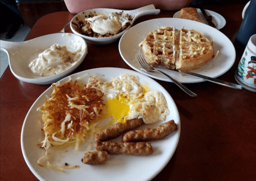 A table with plates of breakfast food: eggs, hash browns, sausage, waffles, and a side of gravy.