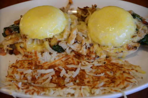 A plate of eggs Benedict with spinach and chicken, topped with hollandaise sauce, served with crispy hash browns.