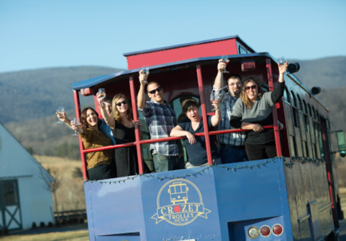 A group of seven people cheer from a blue trolley, holding glasses, with mountains in the background on a sunny day.