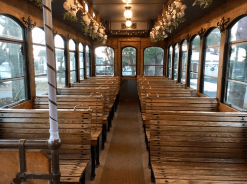 Interior of a vintage trolley with wooden benches and floral decorations, featuring large windows and warm lighting.