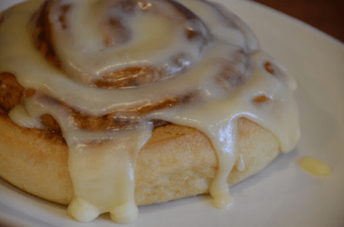 A close-up of a cinnamon roll topped with creamy icing, resting on a white plate.