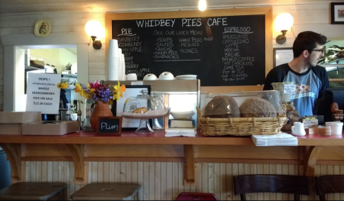 A cozy café counter with baked goods, flowers, and a chalkboard menu listing pies and drinks. A barista is behind the counter.