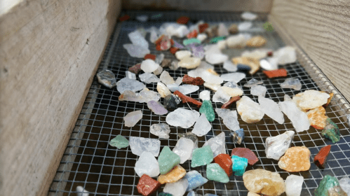 A collection of colorful gemstones and crystals scattered on a mesh screen inside a wooden box.