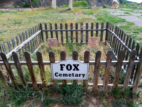 A wooden fenced area labeled "Fox Cemetery" with overgrown grass and a few markers inside.